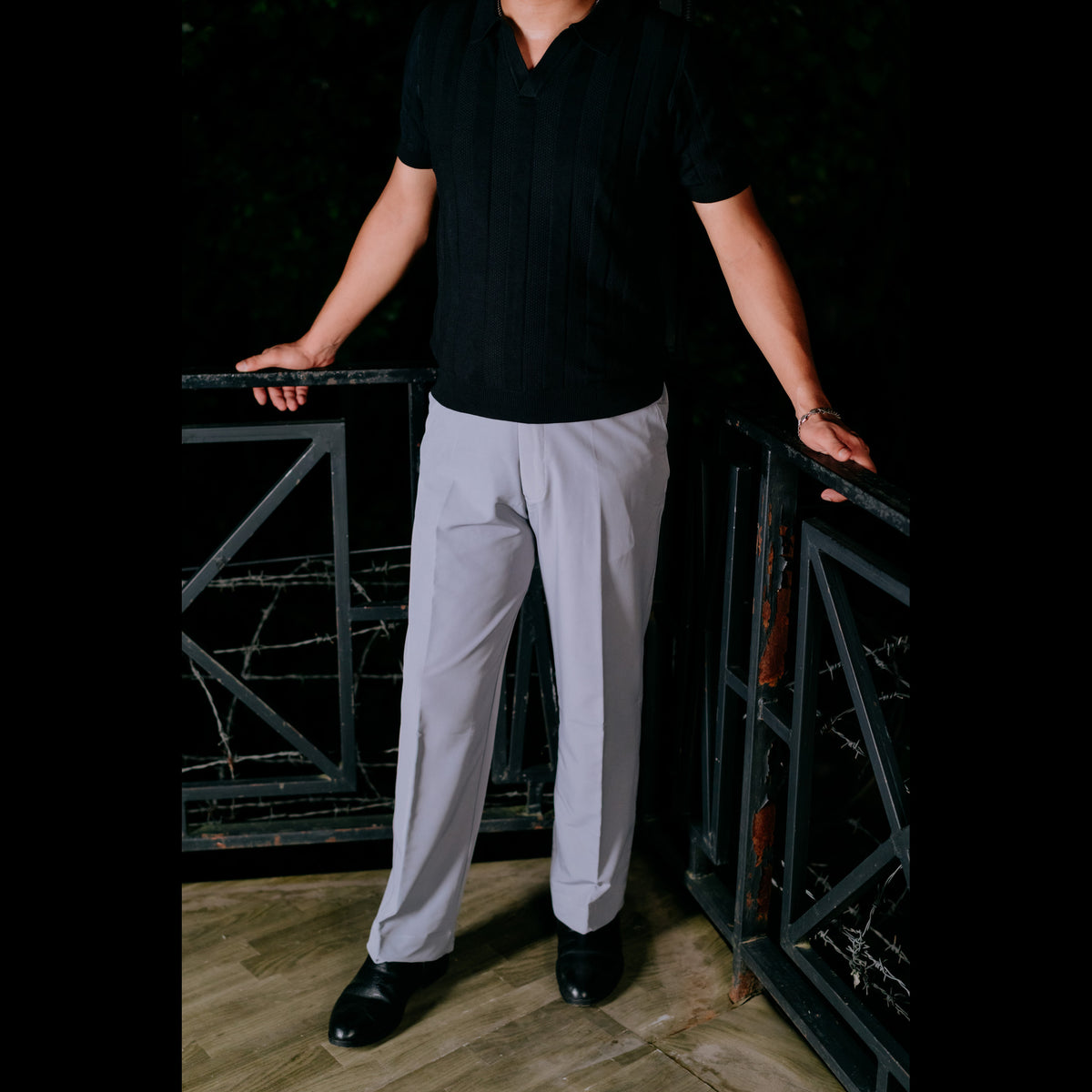 A man wearing light gray dress pants, a black short-sleeve polo shirt, and black dress shoes, standing on a wooden deck between black metal gates at night in NAJRAAN