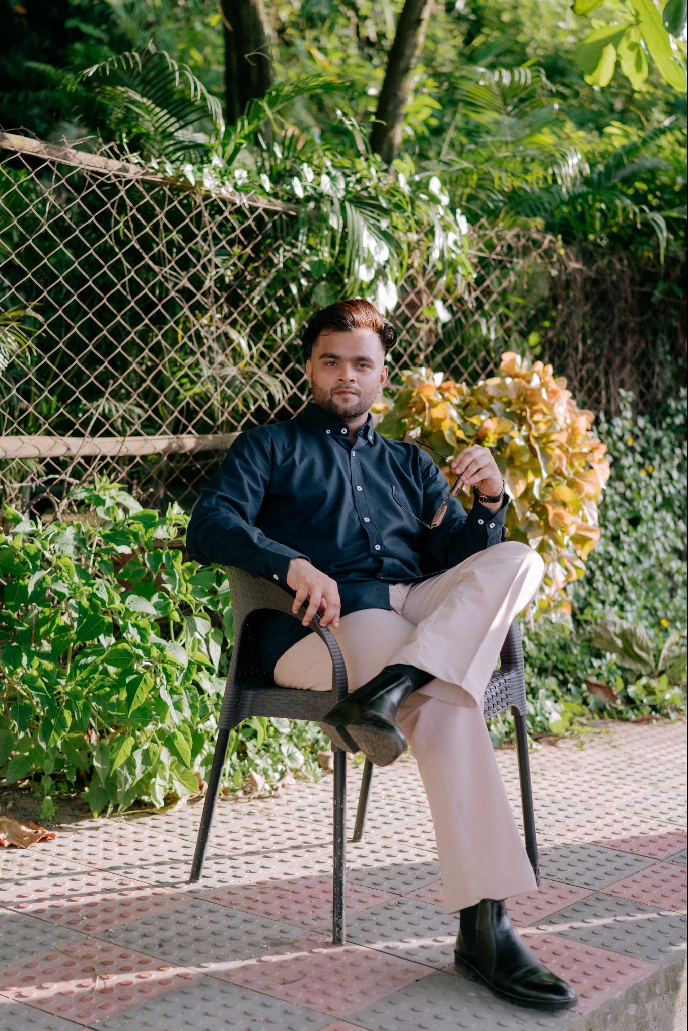 A man wearing a dark navy blue button-up shirt and light beige trousers sits on a black outdoor chair in a garden setting with a chain-link fence and green foliage behind him.