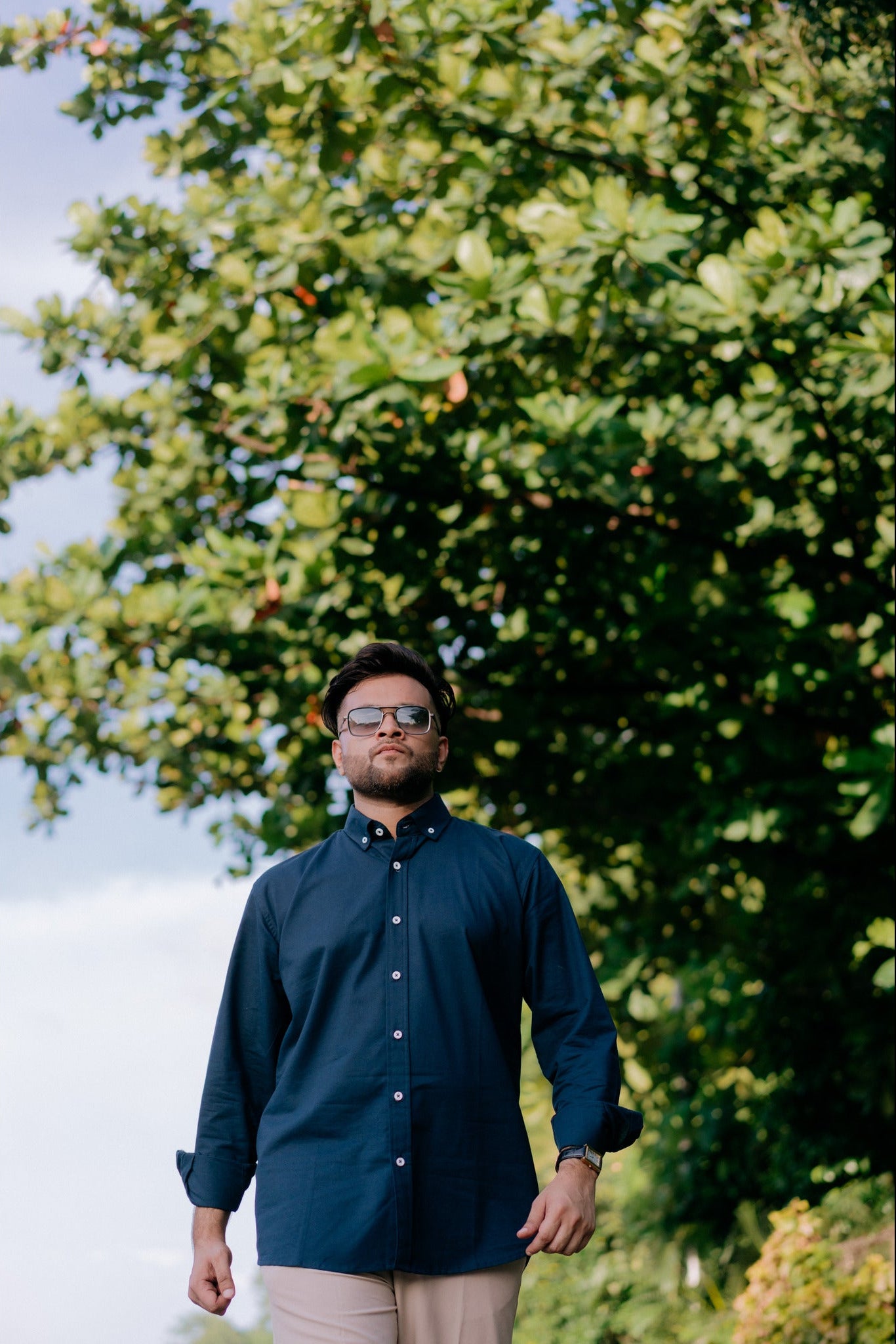 A man wearing a dark navy blue button-down shirt and beige trousers stands outdoors in front of a large green tree.