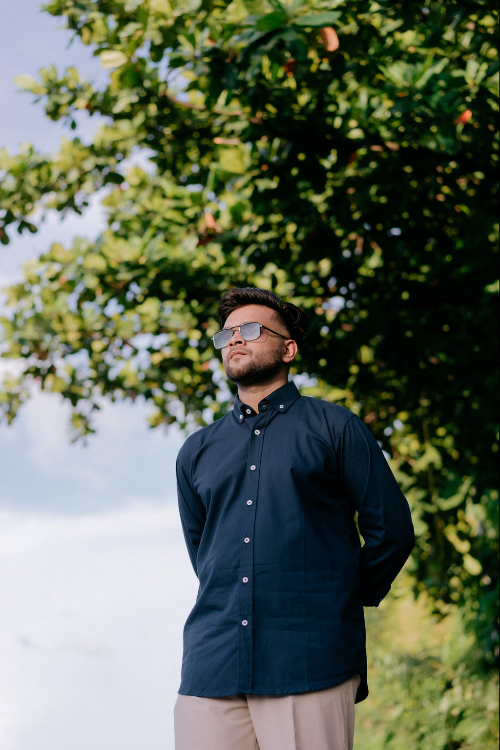 A man wearing a dark navy blue long-sleeve button-down shirt and beige trousers stands outdoors under a leafy green tree, looking upward. He is wearing sunglasses with a gold frame.