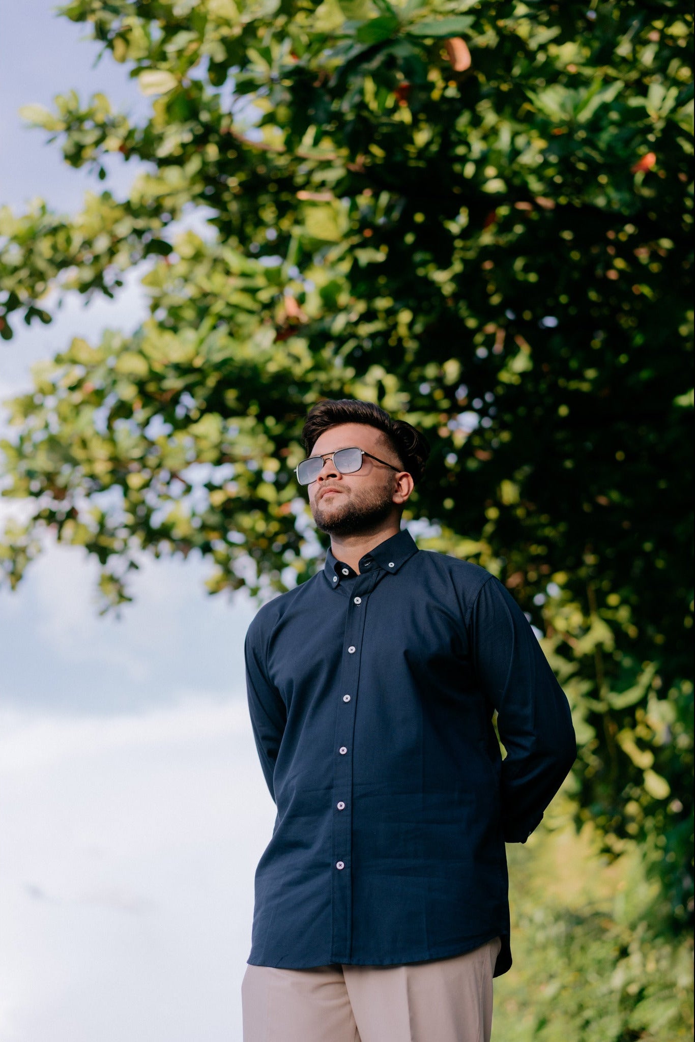 A man wearing a dark navy blue long-sleeve button-down shirt and beige trousers stands outdoors under a leafy green tree, looking upward. He is wearing sunglasses with a gold frame.