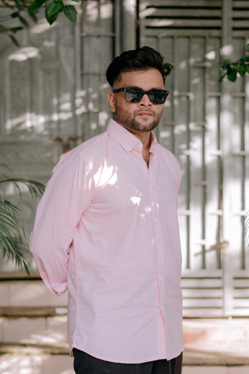 A man wearing a light pink long-sleeve button-up shirt and black sunglasses, standing outdoors in front of a white metal gate with dappled sunlight filtering through leaves.