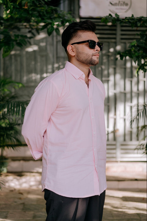 A man wearing a light pink long-sleeve button-down shirt and black trousers, standing outdoors in front of a metal gate with green foliage.