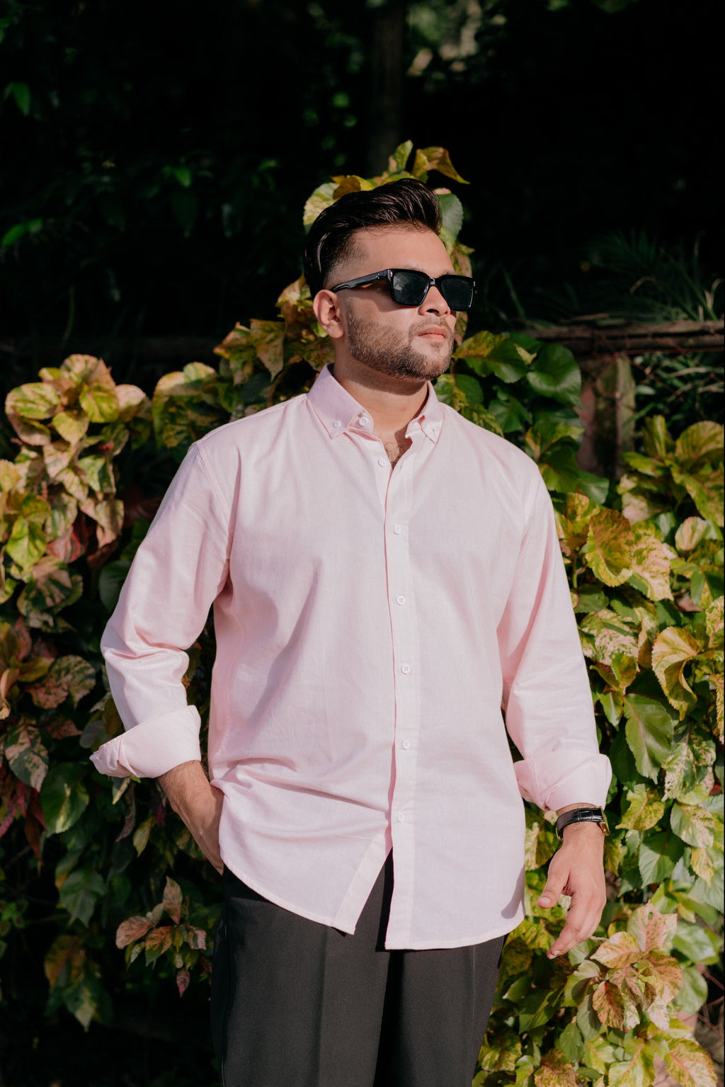 A man wearing a light pink long-sleeve button-down shirt and dark trousers, standing outdoors in front of a leafy green hedge.