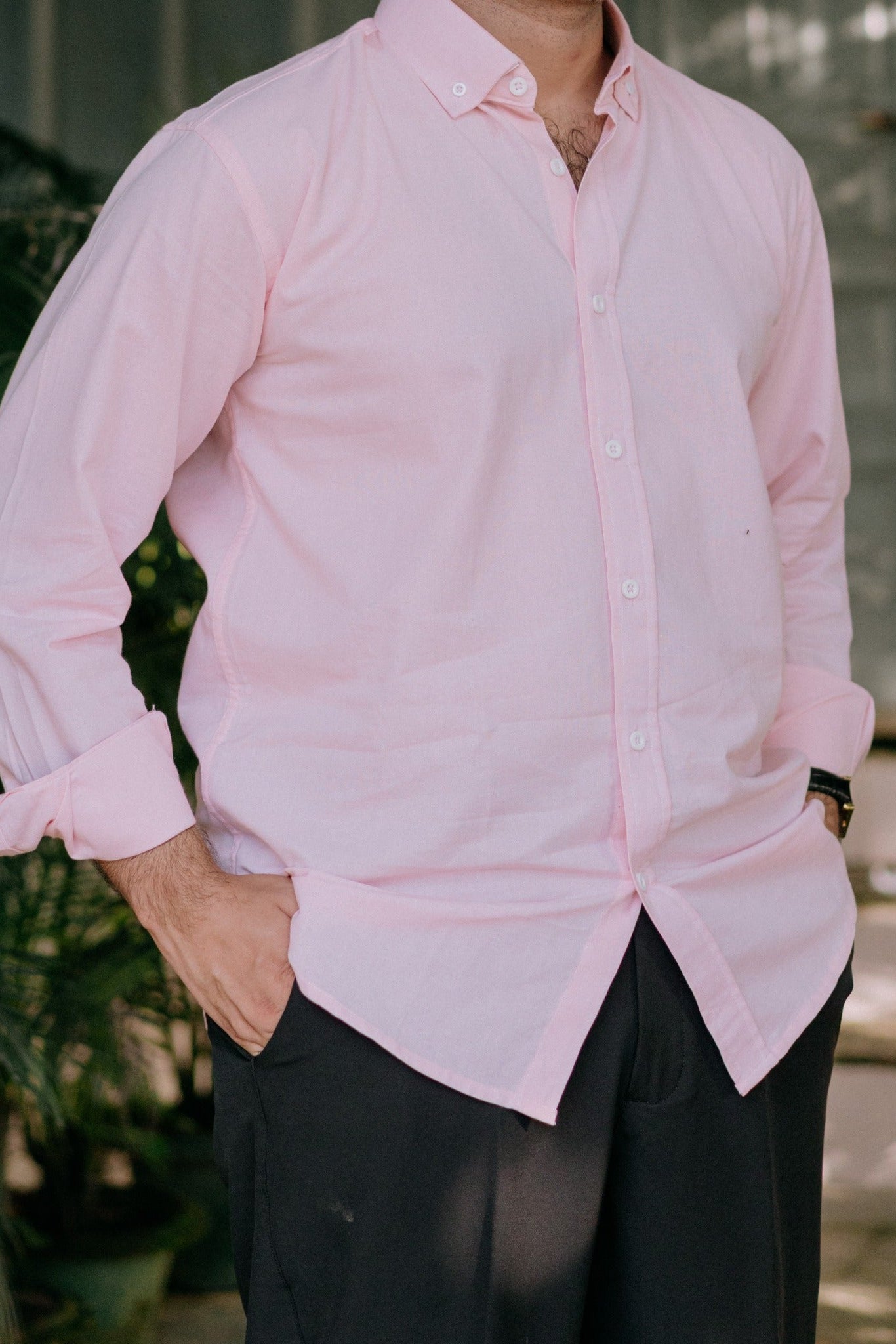 A man wearing a light pink long-sleeve button-down shirt with black trousers, standing in a softly blurred indoor/outdoor setting with greenery in the background.