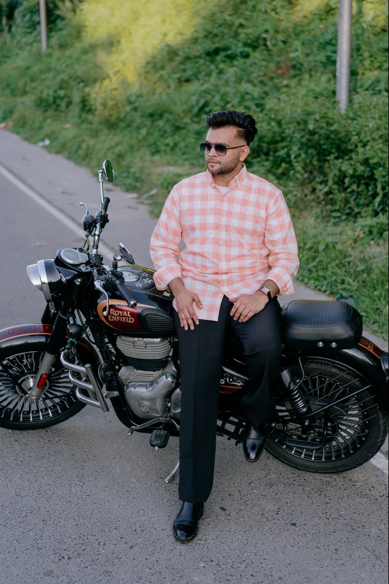 A man wearing a pink and white checkered shirt, black trousers, and sunglasses sits on a black Royal Enfield Classic Cruiser motorcycle on a paved road with green foliage in the background.