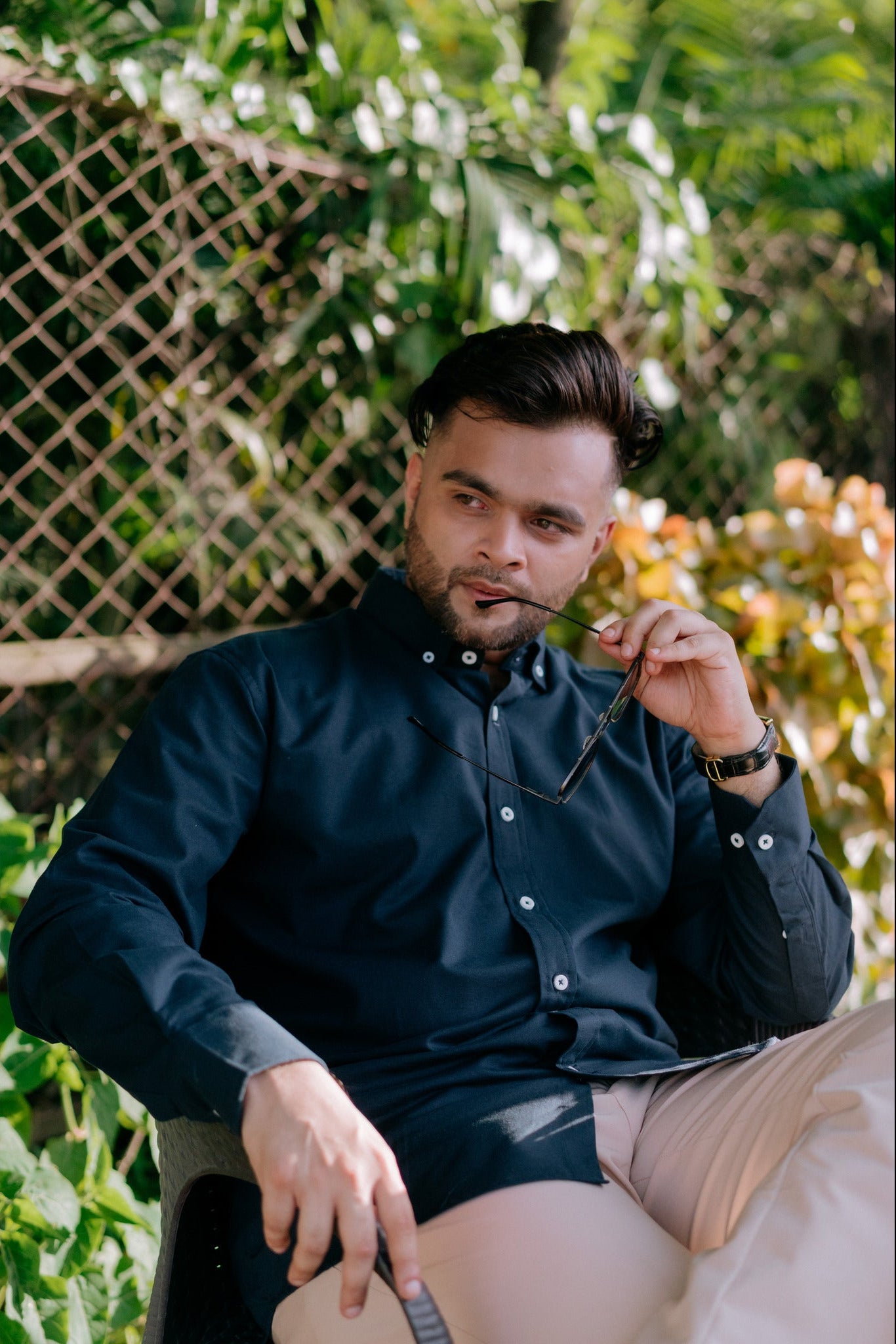 A man wearing a dark navy blue long-sleeve button-up shirt and beige trousers, sitting outdoors in front of a green lattice fence.