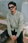 A man wearing black sunglasses, a light sage green button-down shirt, and black trousers, sitting on stone steps against a light-colored wall.