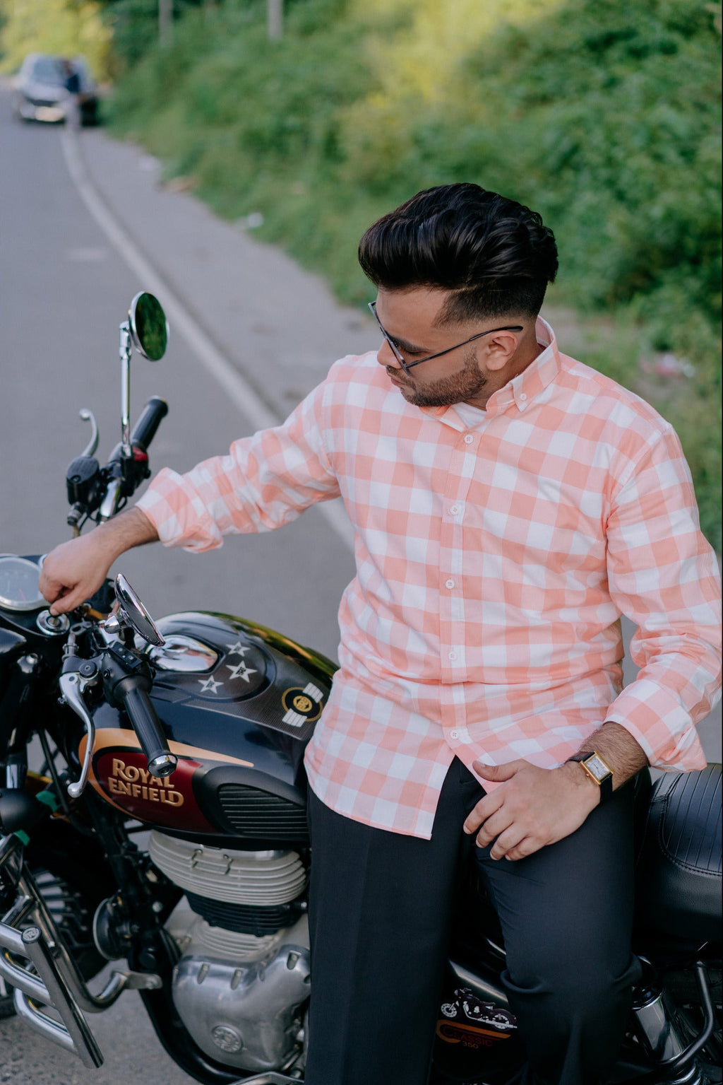 A man wearing a pink and white checkered long-sleeve shirt and dark trousers sits on a black Royal Enfield motorcycle on a paved road with green foliage in the background.