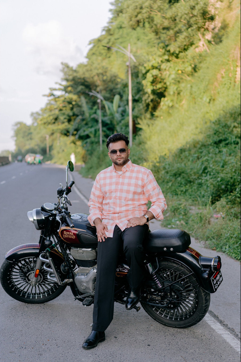 A man wearing a pink and white checkered shirt and black trousers sits on a black cruiser motorcycle on a paved road, with a lush green hillside in the background.