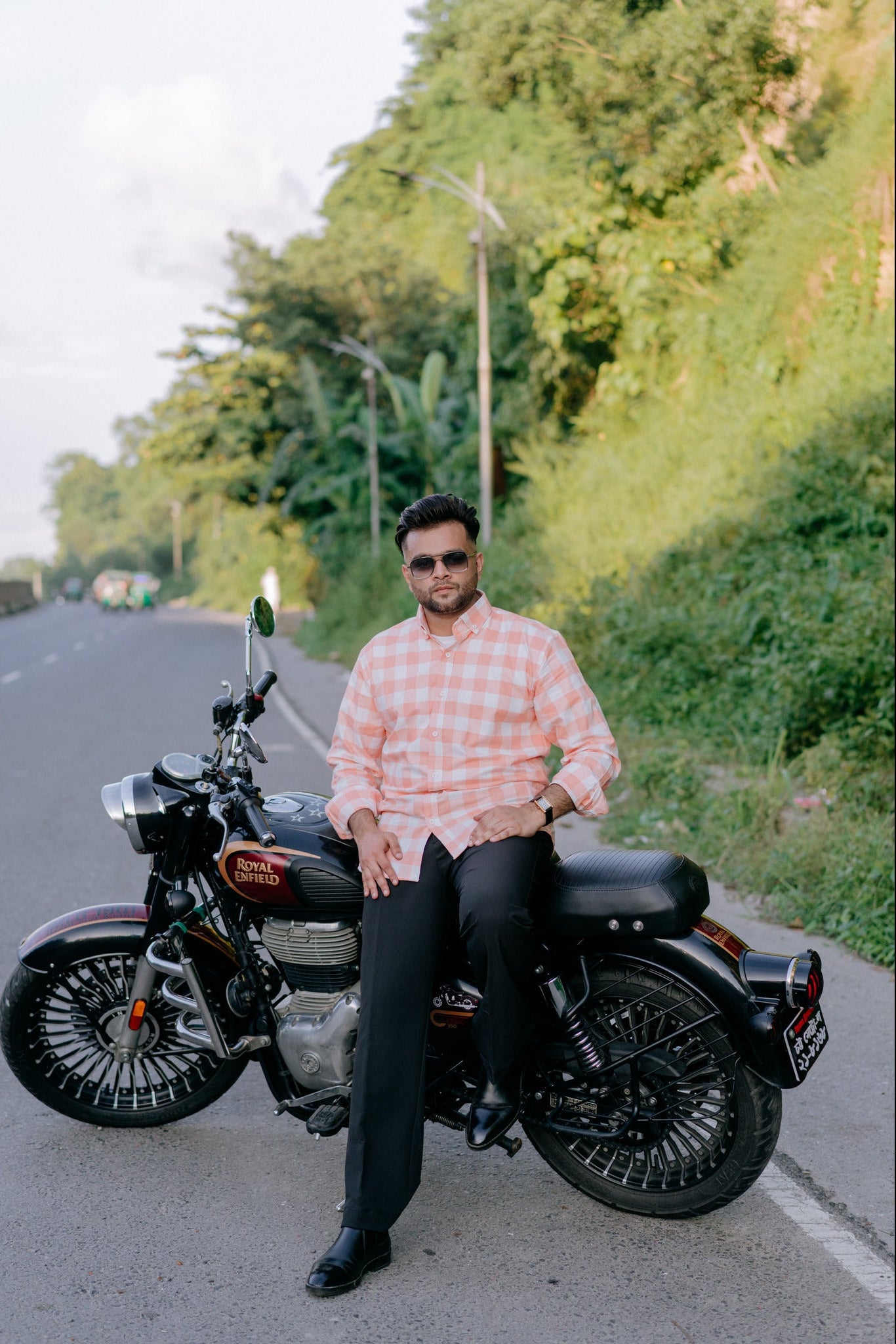 A man wearing a pink and white checkered shirt and black trousers sits on a black cruiser motorcycle on a paved road, with a lush green hillside in the background.