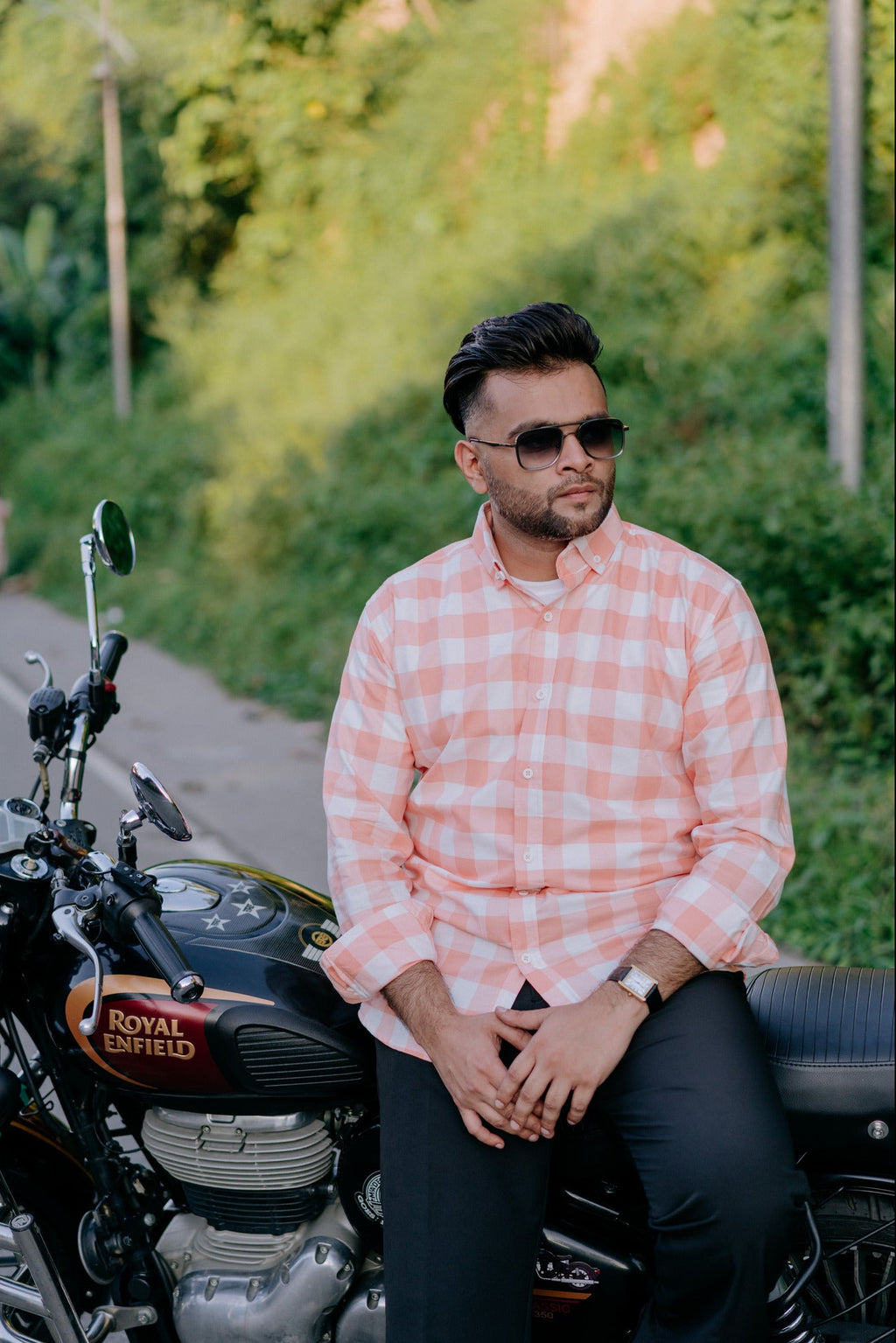 A man wearing a pink and white checkered button-down shirt and sunglasses sits on a black Royal Enfield motorcycle on a paved path surrounded by green foliage.