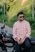 A man wearing a pink and white checkered button-down shirt and sunglasses sits on a black Royal Enfield motorcycle on a paved path surrounded by green foliage.