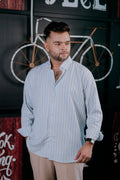 A man wearing a light blue and white vertically striped long-sleeve button-down shirt with beige trousers, standing in front of a dark wall with a bicycle graphic.