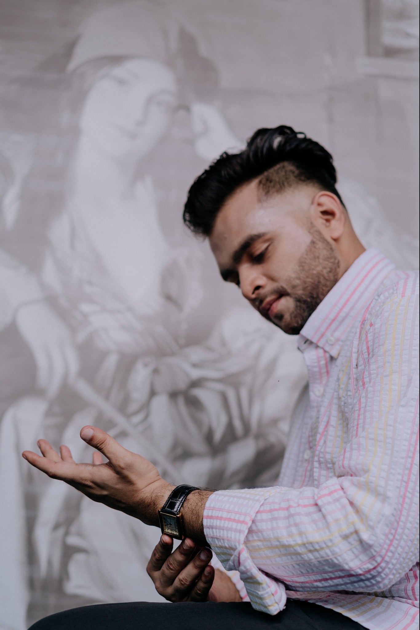 A man in a light pink and white striped button-down shirt is shown in profile, looking down while wearing a black smartwatch. The background is a large, desaturated black-and-white sketch of a woman.