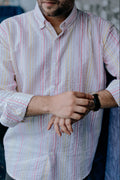 A man wearing a white long-sleeve button-down shirt with vertical pink, yellow, and blue stripes, shown from the chin down as he adjusts his watch.