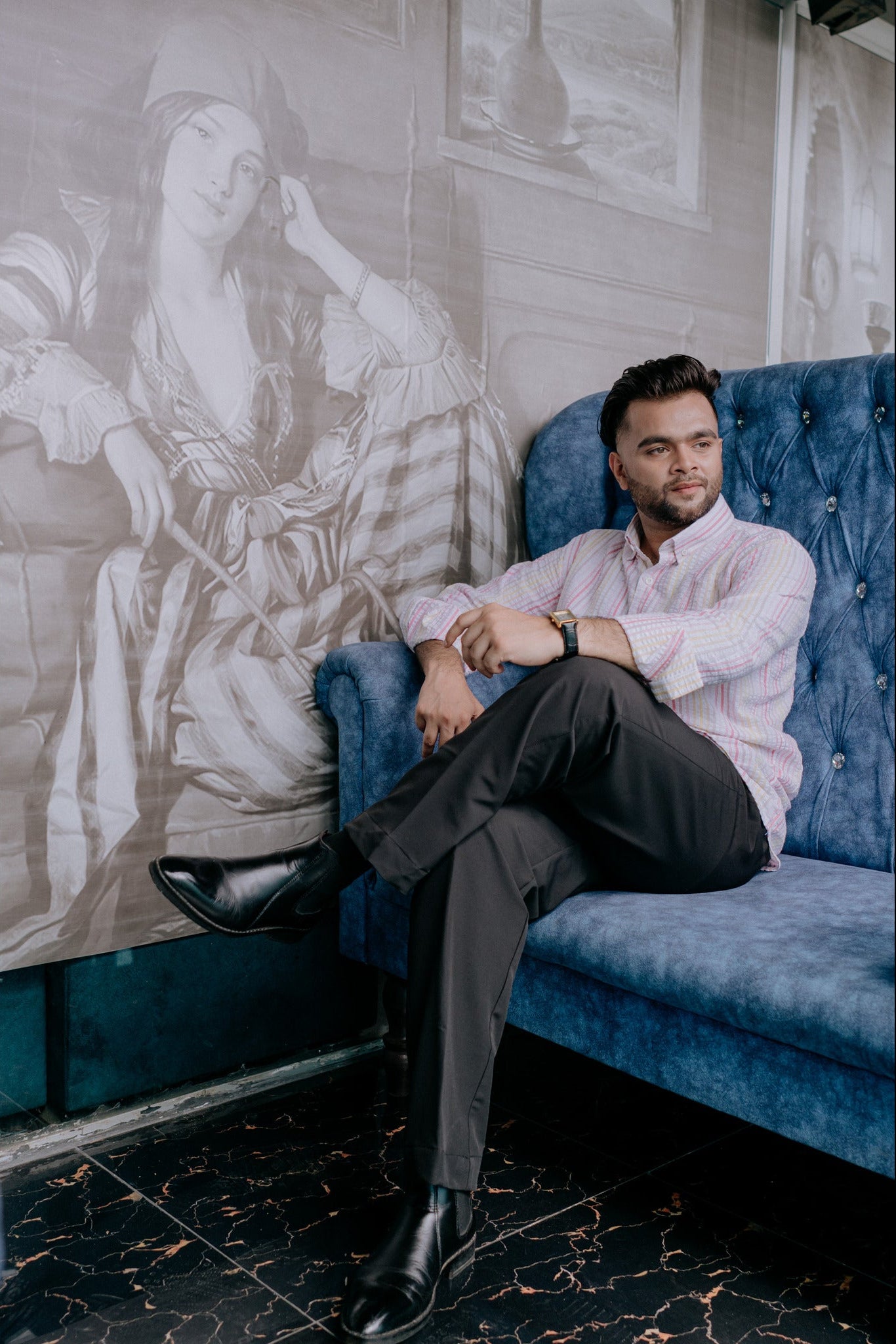 A man wearing a light pink striped button-down shirt, dark gray trousers, and black dress shoes sits on a blue tufted velvet sofa in front of a large grayscale classical painting.