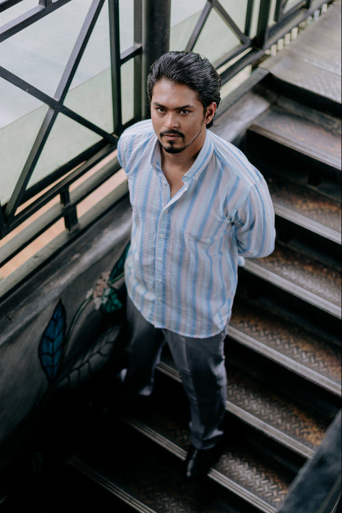 A man wearing a light blue and white vertically striped button-down shirt and dark gray trousers, standing on an outdoor staircase.