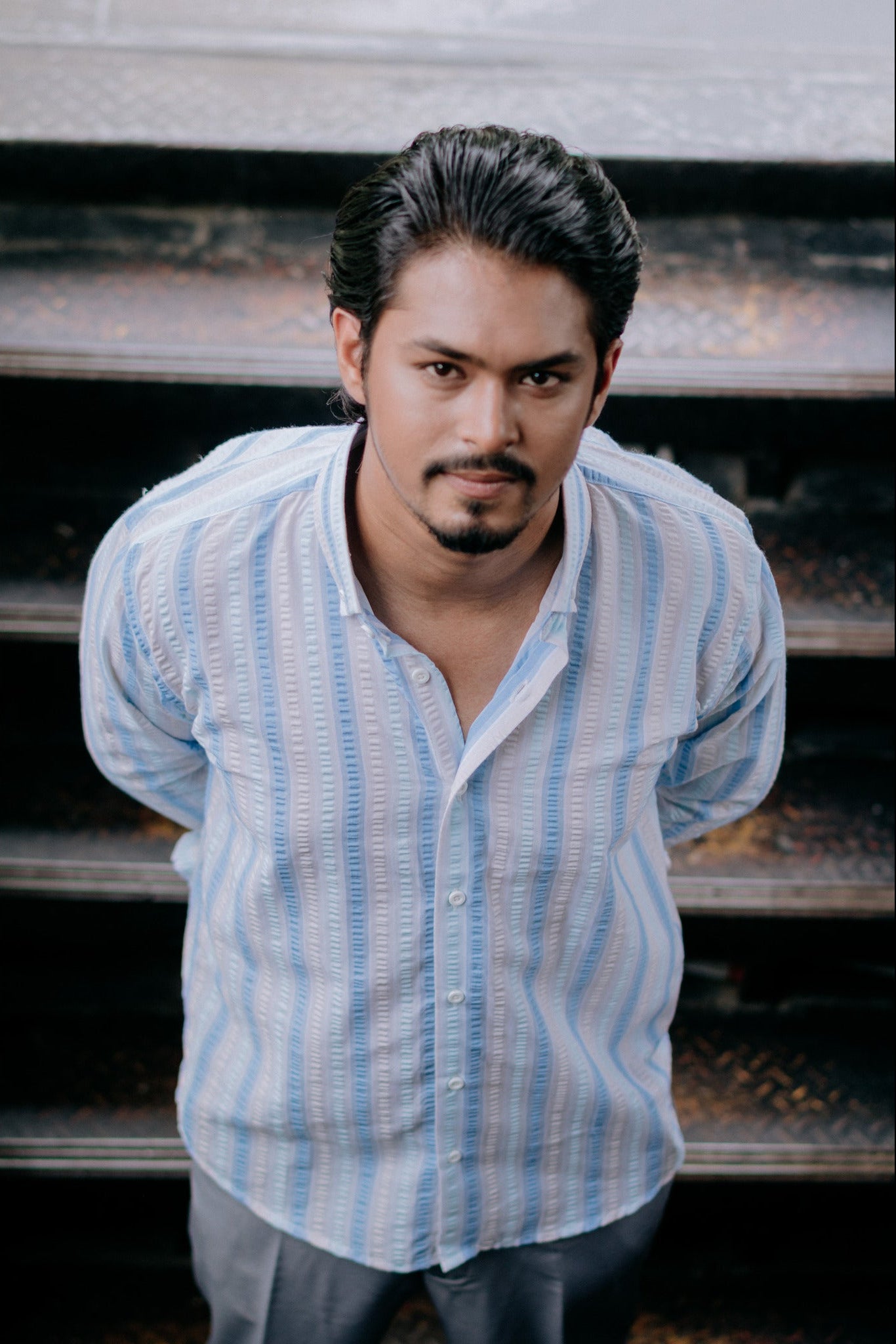 A man wearing a light blue and white vertically striped button-down shirt, standing in front of dark outdoor steps.