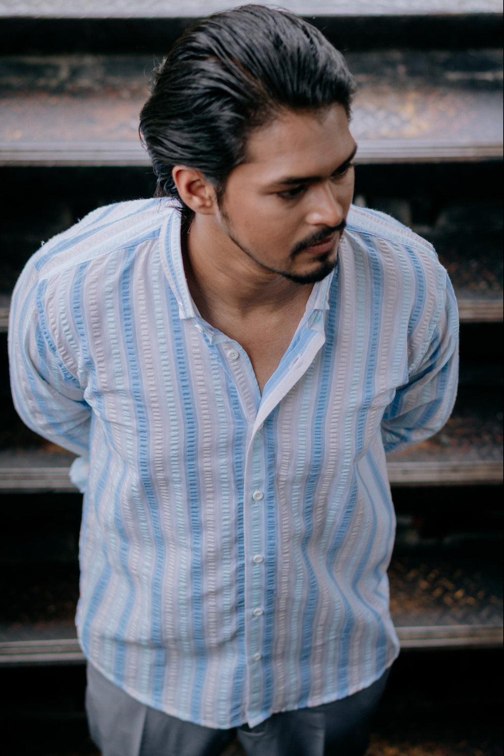 A man wearing a light blue and white vertically striped button-up shirt, standing in front of dark steps.
