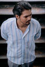A man wearing a light blue and white vertically striped button-up shirt, standing in front of dark steps.