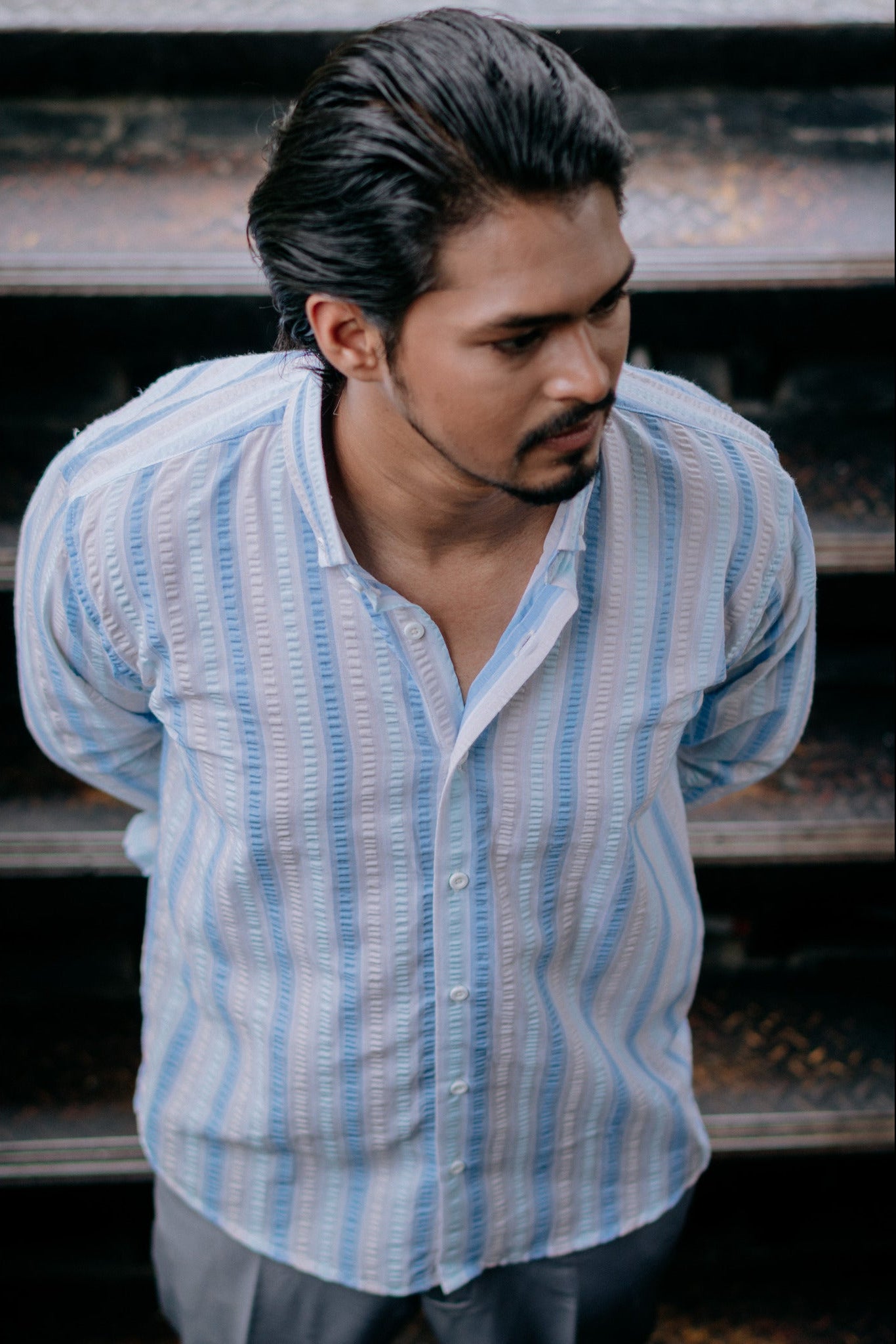 A man wearing a light blue and white vertically striped button-up shirt, standing in front of dark steps.
