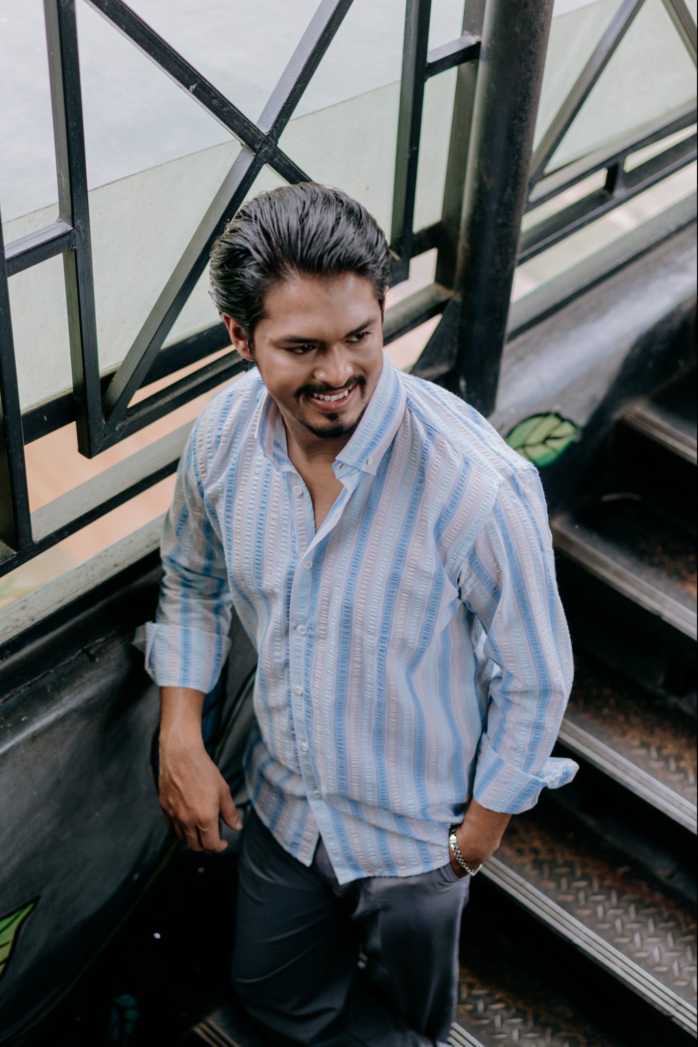 A man wearing a light blue and white vertically striped button-down shirt and dark gray trousers, smiling as he stands on metal stairs next to a glass-paneled wall.