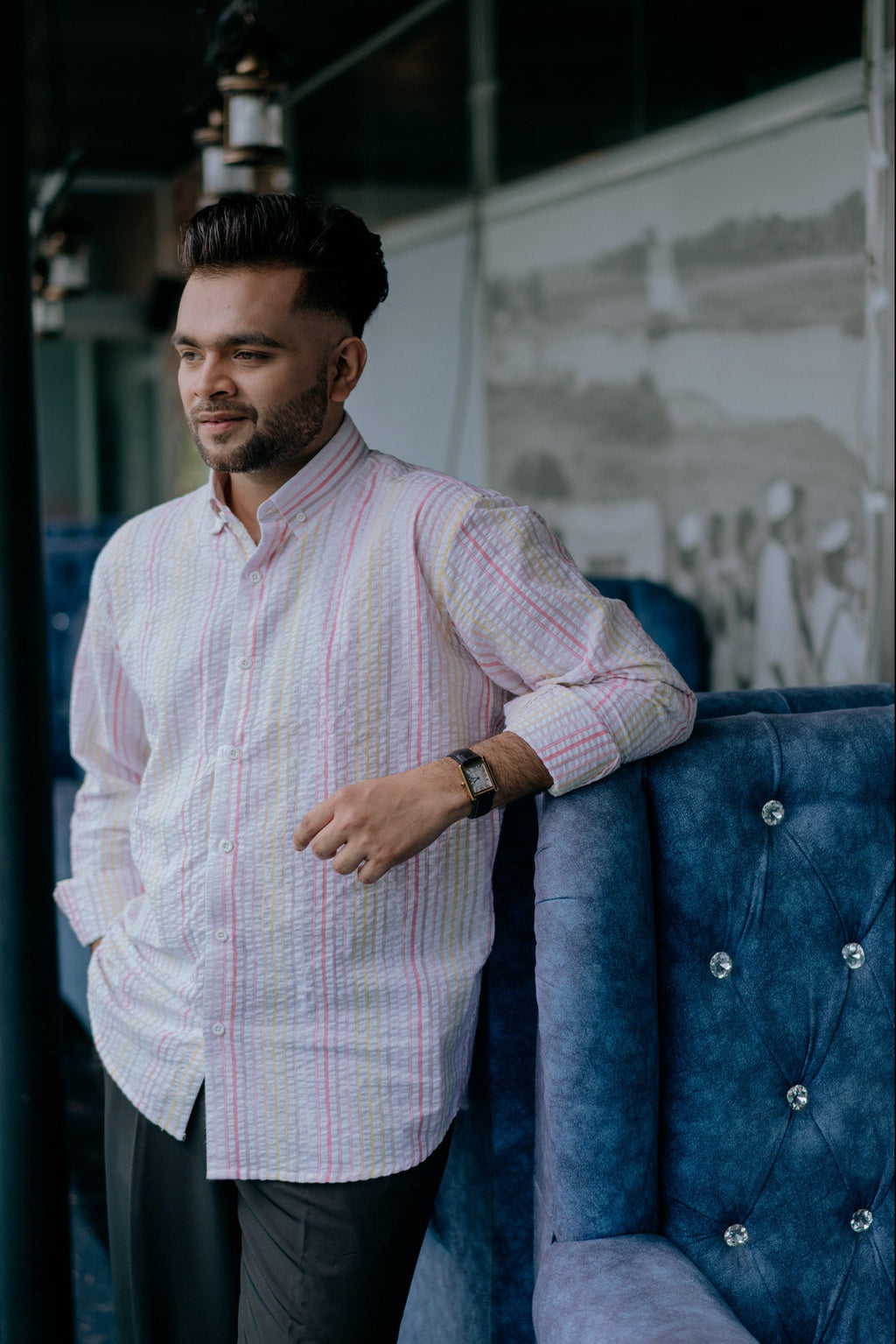 A man wearing a light pink and white vertically striped long-sleeve button-up shirt leans against a blue tufted armchair in an indoor setting.