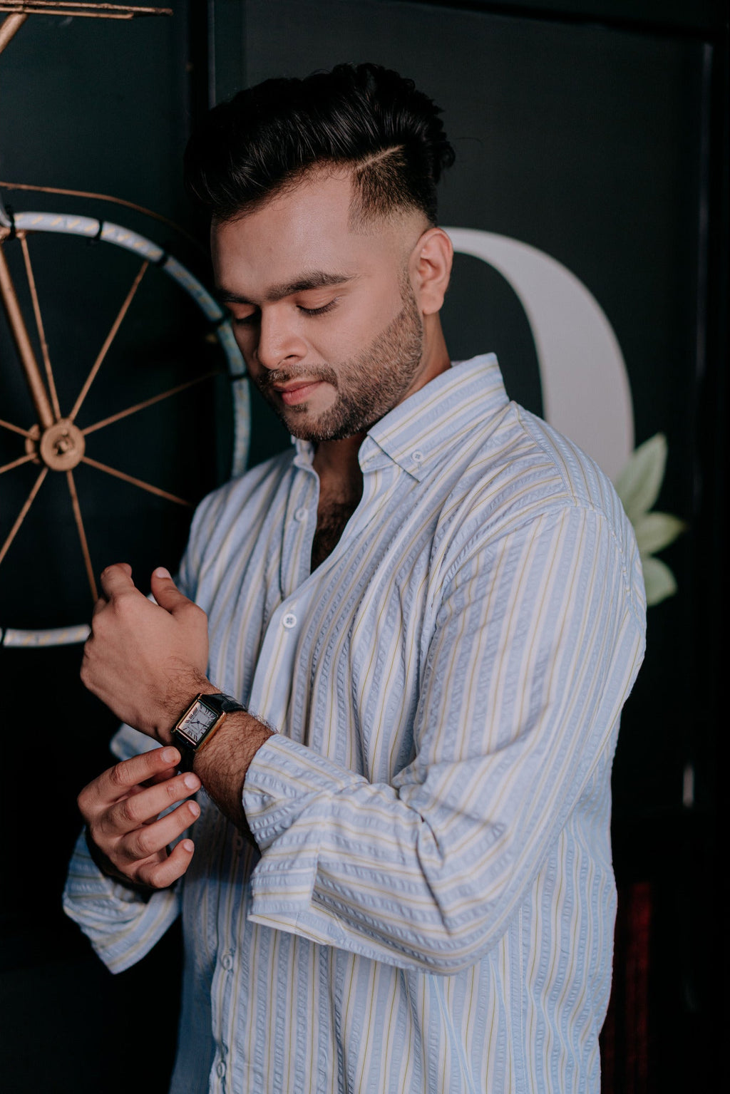 A man wearing a light blue and white vertically striped button-down shirt, looking down while adjusting his black wristwatch.