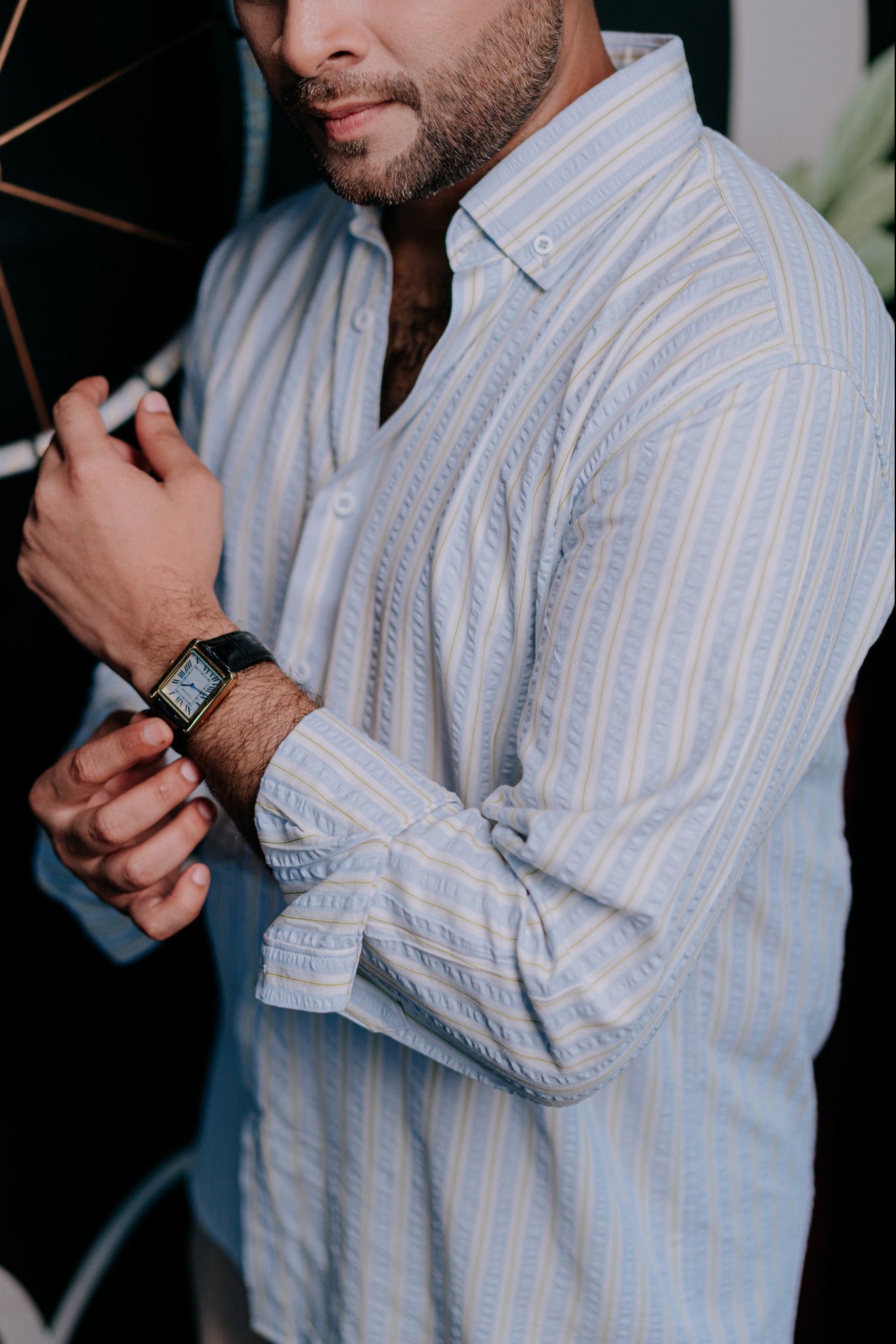 A man wearing a light blue and white vertically striped button-down shirt, shown from the chest down as he adjusts his watch.