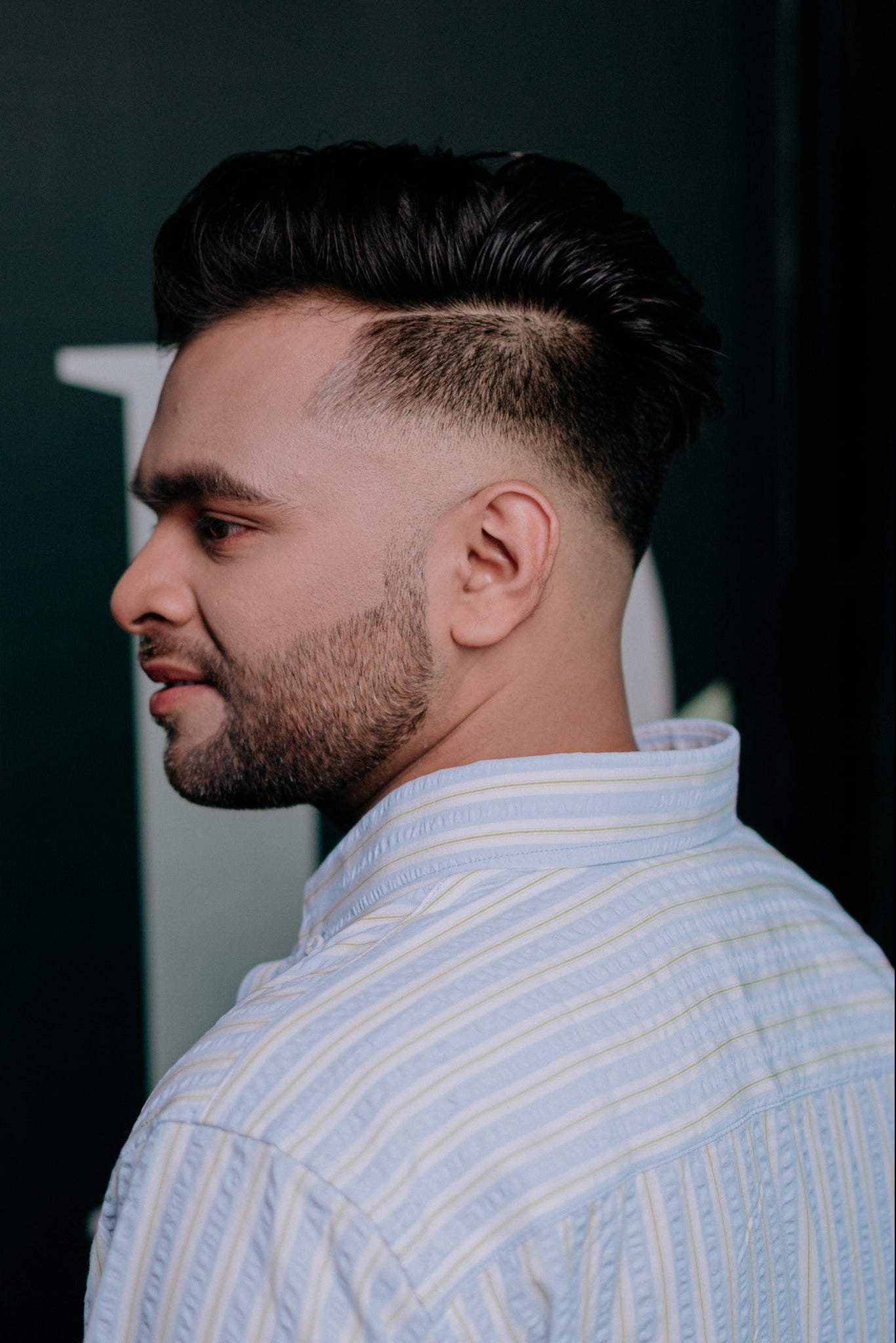 A man in profile wearing a light blue and white vertically striped button-down shirt, showcasing a modern fade haircut.
