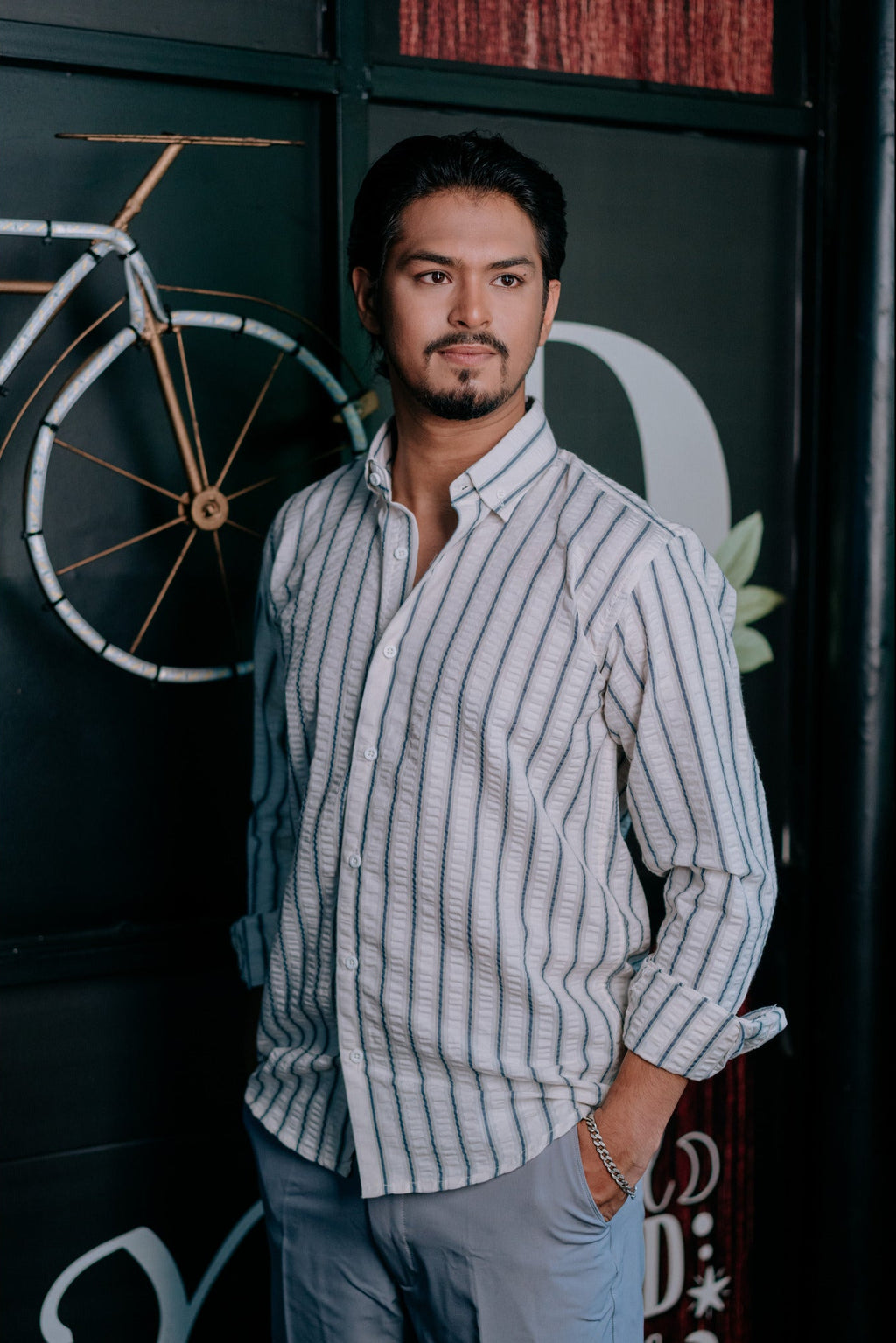 A man wearing a light gray and white vertically striped button-down shirt and light gray trousers, standing in front of a dark wall with a bicycle graphic.