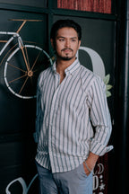 A man wearing a light gray and white vertically striped button-down shirt and light gray trousers, standing in front of a dark wall with a bicycle graphic.