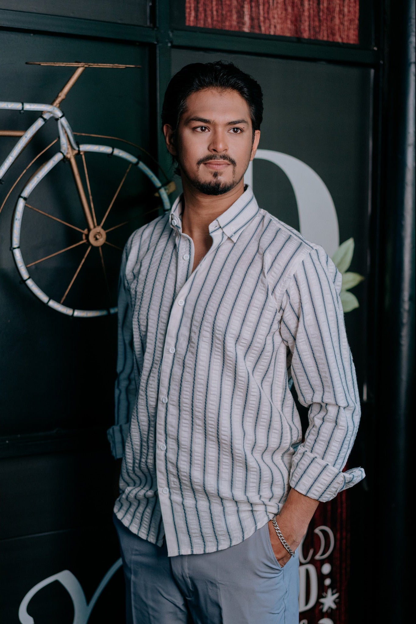 A man wearing a light gray and white vertically striped button-down shirt and light gray trousers, standing in front of a dark wall with a bicycle graphic.