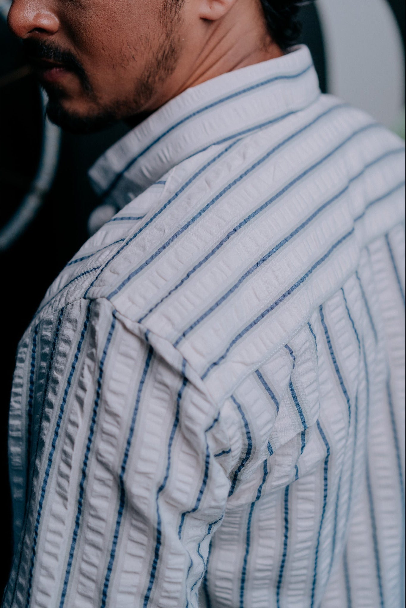 Close-up side view of a man wearing a textured white shirt with thin vertical navy stripes, showing the fabric's crinkled texture and collar.