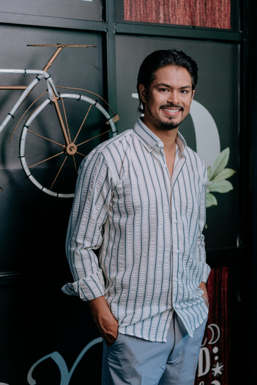 A man wearing a light blue and white vertically striped button-down shirt and light blue trousers, standing in front of a dark wall with a bicycle decoration.