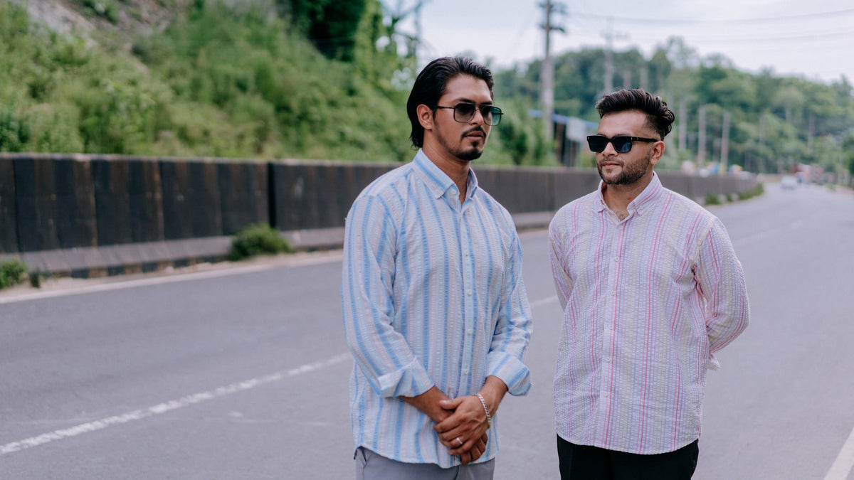 Two men standing on a road outdoors, wearing light-colored striped button-up shirts. One shirt is light blue and white, and the other is white with subtle pink and blue stripes.